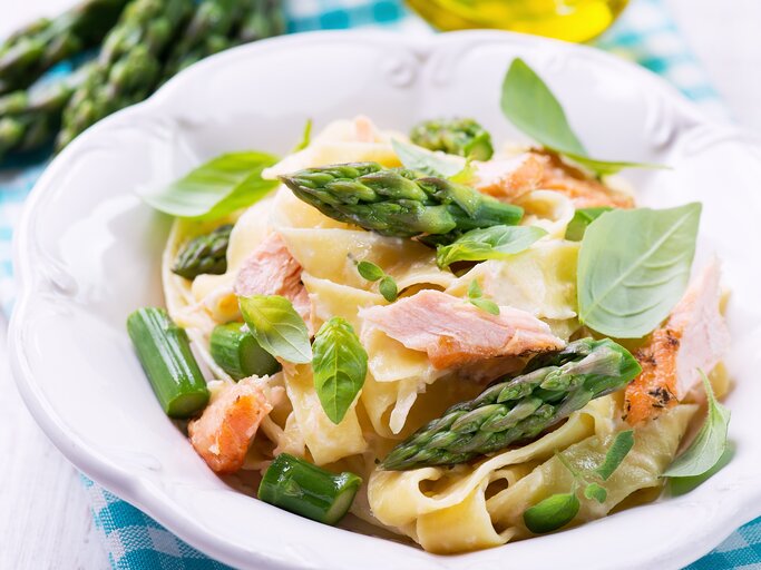 Ein weißer Teller mit dem Pastagericht auf einem hellen Holztisch mit blau-weiß-karierter Decke. Hinten unscharf Olivenöl und grüner Spargel zu erkennen. | © Shutterstock/ Kamila i Wojtek Cyganek
