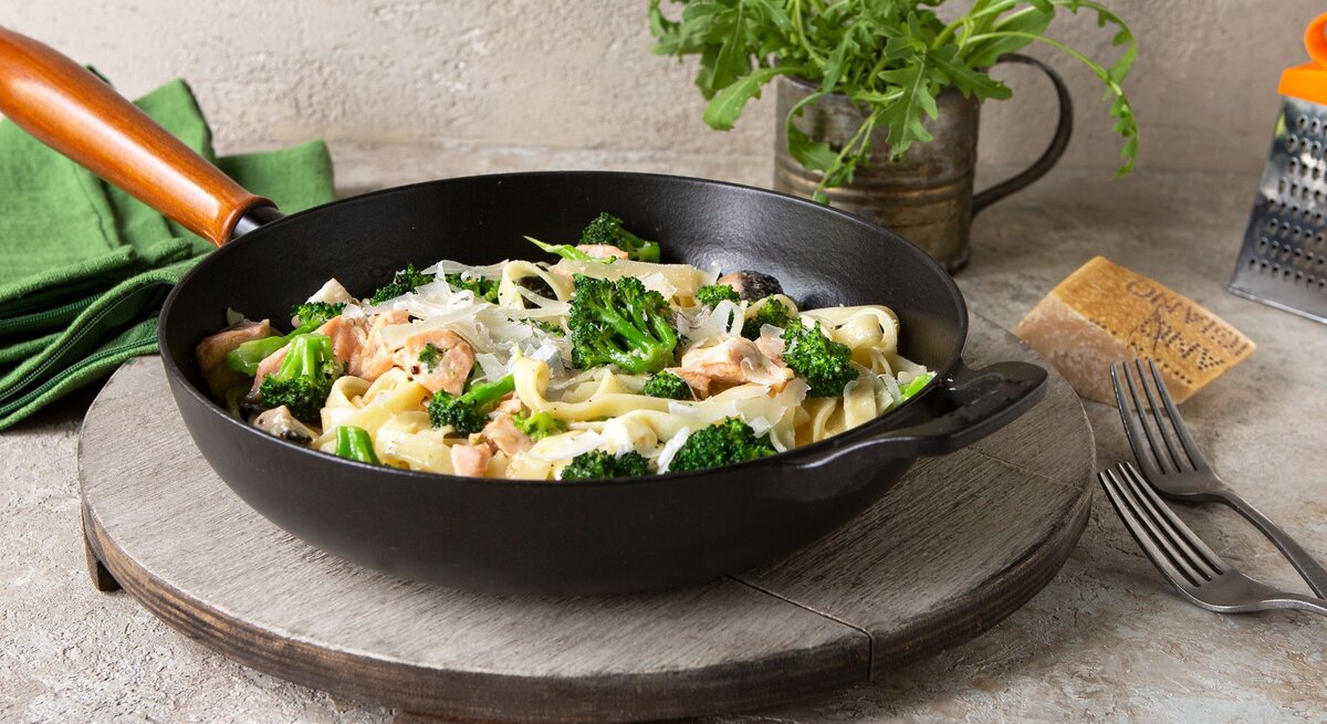 Eine schwarze Pfanne mit Tagliatelle mit Lachs und Brokkoki auf einer grauen Scheibe.