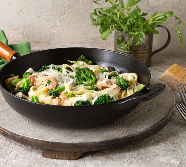 Eine schwarze Pfanne mit Tagliatelle mit Lachs und Brokkoki auf einer grauen Scheibe. | © AdobeStock/kochabamba