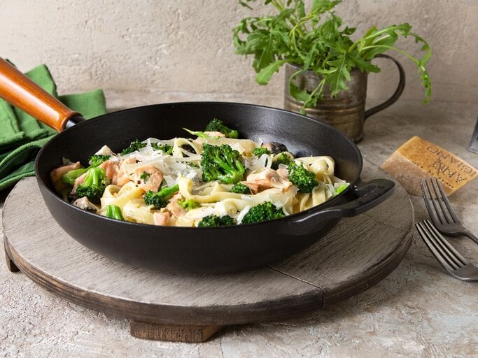 Eine schwarze Pfanne mit Tagliatelle mit Lachs und Brokkoki auf einer grauen Scheibe. | © AdobeStock/kochabamba