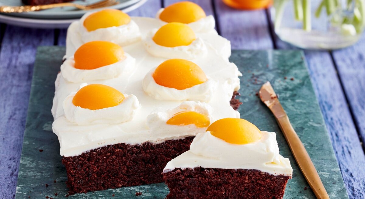 Auf einer Platte liegen Spiegeleier-Brownies. Sie sind mit Sprikosenhälften und einer Ricotta-Creme dekoriert und sehen aus wie aufgeschlagene Eier. 