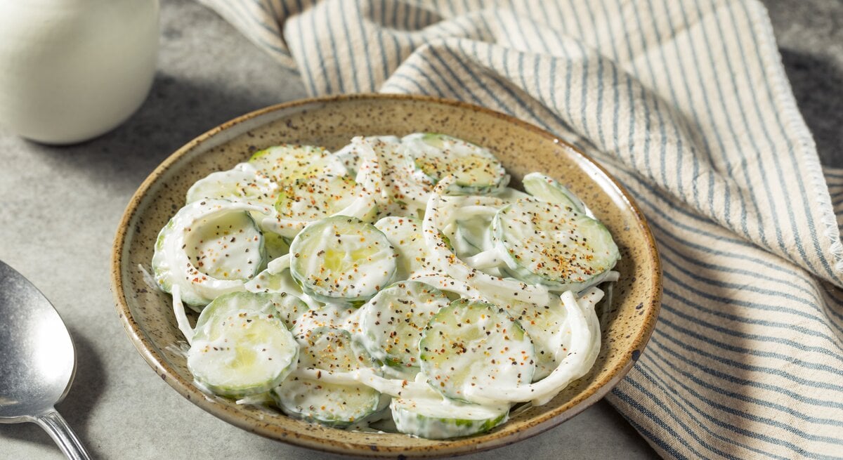 Sauerrahm-Gurkensalat mit frischem Pfeffer neben Geschirrtuch auf grauem Untergrund.