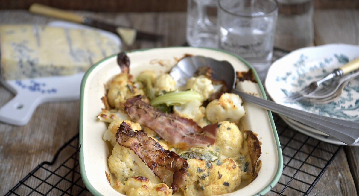 Blumenkohlgratin mit Blauschimmelkaese und gerösteten Speckstreifen, serviert in einer Auflaufform auf einem Rost auf einem gedeckten Holztisch. 