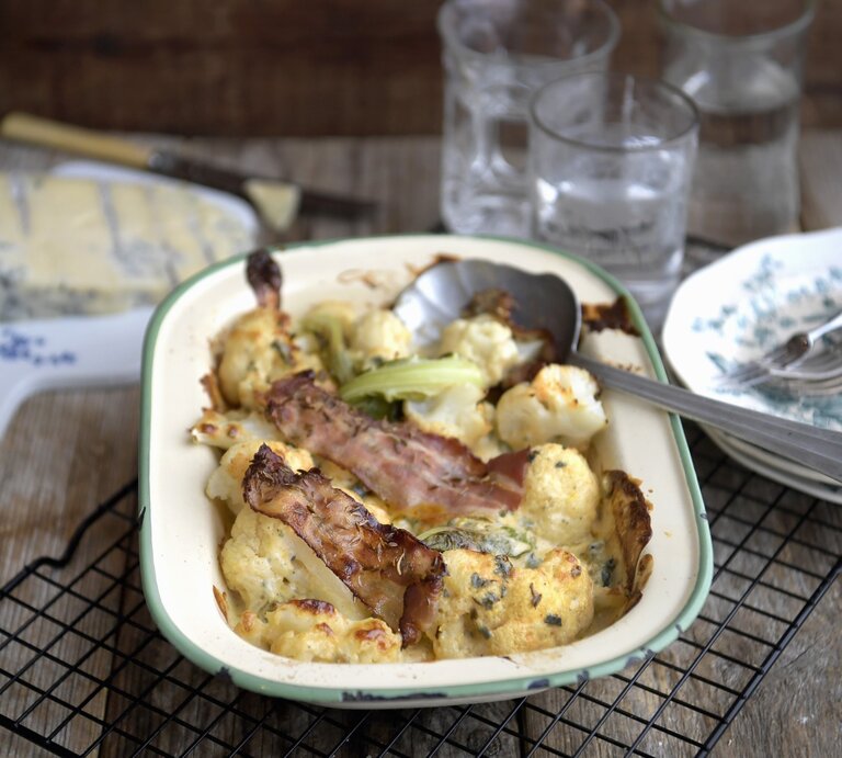 Blumenkohlgratin mit Blauschimmelkaese und gerösteten Speckstreifen, serviert in einer Auflaufform auf einem Rost auf einem gedeckten Holztisch.  | © StockFood / TT News Agency / TT News Agency