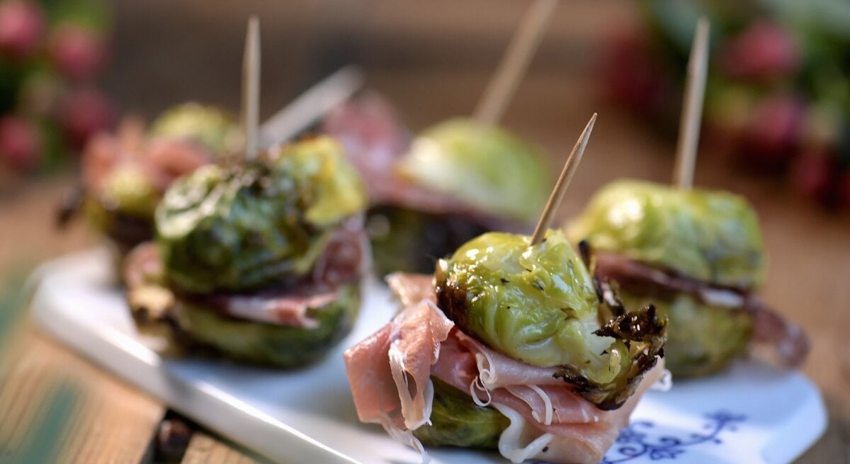 Rosenkohl-Spießchen mit Salami und Prosciutto auf einem weißen Brett mit unscharfem Hintergrund.