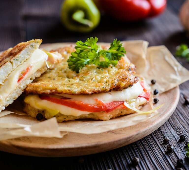 Die Low-Carb-Panini mit Mozzarella und Tomate auf einem Brett. Dahinter verschiedene Zutaten auf einem hölzernen Untergrund. | © Getty Images / NoirChocolate