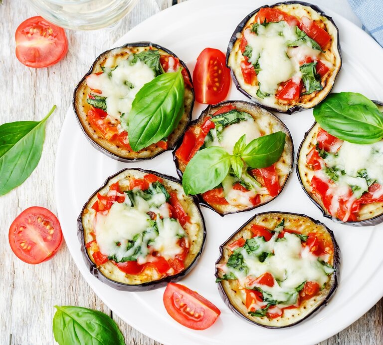 Mehrere Auberginen-Pizzen auf einem weißen Teller und hellem Holztisch von oben fotografiert. | © Shutterstock/ Nataliya Arzamasova