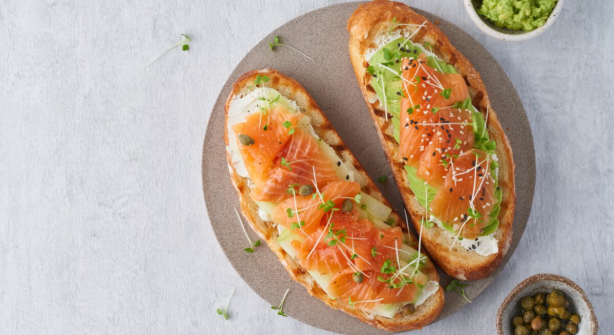 Draufsicht: Ein Teller auf dem zwei Low-Carb-Toast mit Lachs liegen. Daneben ist ein rundes Gefäß mit Avocadocreme und eines mit Kapern.