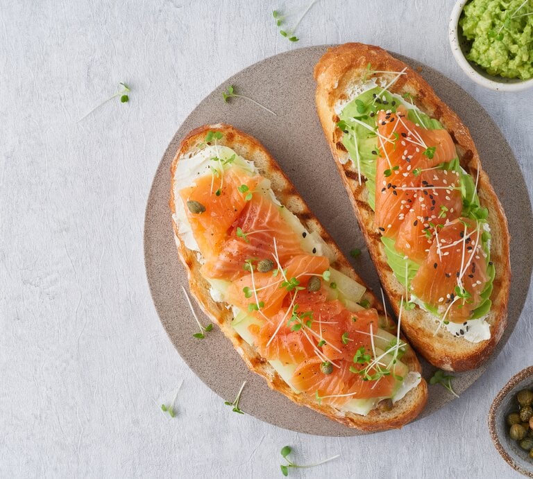 Draufsicht: Ein Teller auf dem zwei Low-Carb-Toast mit Lachs liegen. Daneben ist ein rundes Gefäß mit Avocadocreme und eines mit Kapern. | © Shutterstock / Nata Bene