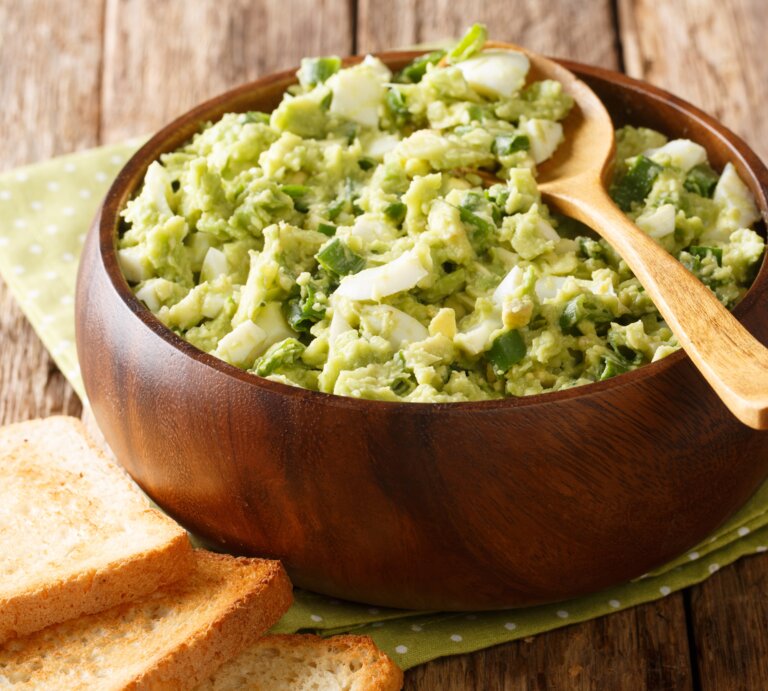 Avocado-Eiersalat mit Lauch und Joghurt in Holzschüssel auf grüner Serviette und Holztisch. Oben auf Holzlöffel. Links am Bildrand geröstetes Toastbrot. Draufsicht. | © Shutterstock / AS Food studio
