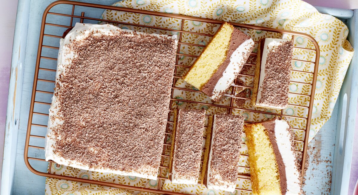 Auf einem Backblech liegt ein eckiges Stück Schoko-Schnitten mit Quark. Im Anschnitt sieht man die verschiedenen Schichten. Oben drauf sind Schokoraspeln. 