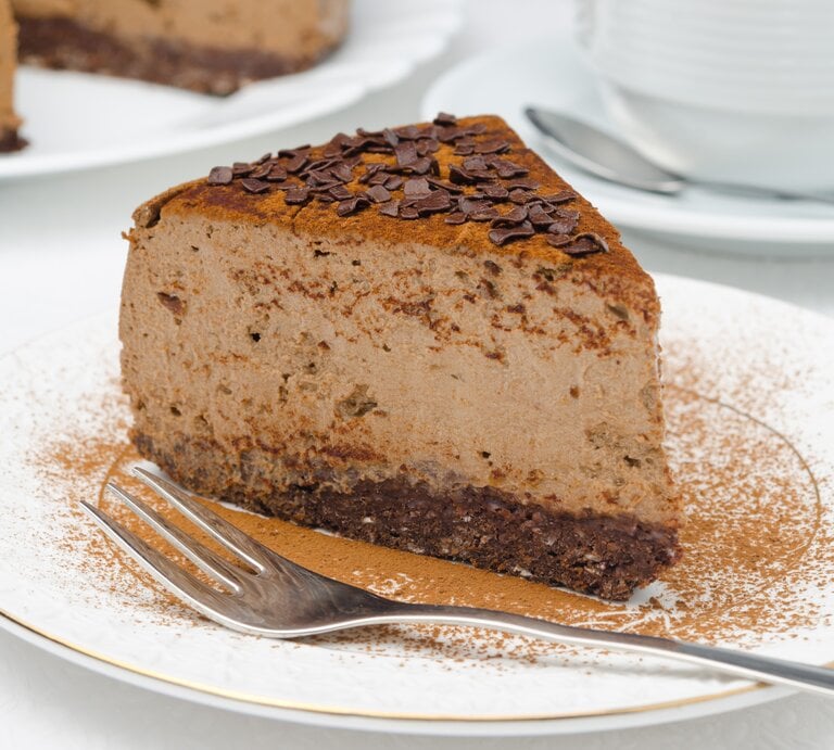 Ein Stück veganer Schokoladen-Cheesecake mit Kakao und Schokosplittern bestreut auf einem kleinen Teller. | © Shutterstock/ Yulia Davidovich