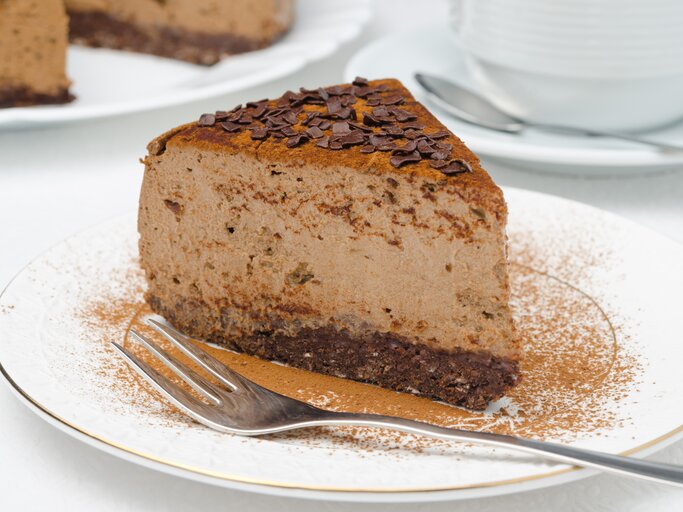 Ein Stück veganer Schokoladen-Cheesecake mit Kakao und Schokosplittern bestreut auf einem kleinen Teller. | © Shutterstock/ Yulia Davidovich