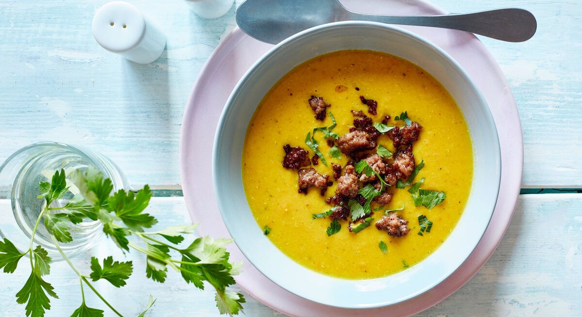 Auf einem Untergrund aus Holz stehen ein Salz und ein Pfefferstreuer. Daneben ein Glas mit einem Zweig Petersilie. Und auch ein Teller auf dem eine Suppenschüssel mit einer Currysuppe mit Hackfleisch   steht. Neben der Schüssel liegt ein Löffel.