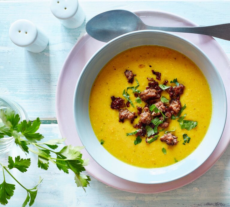 Auf einem Untergrund aus Holz stehen ein Salz und ein Pfefferstreuer. Daneben ein Glas mit einem Zweig Petersilie. Und auch ein Teller auf dem eine Suppenschüssel mit einer Currysuppe mit Hackfleisch   steht. Neben der Schüssel liegt ein Löffel. | © EAT CLUB / Isabell Triemer