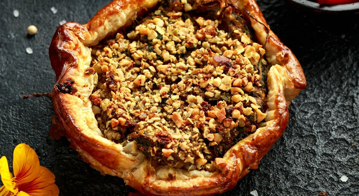 Eine Kürbis-Tarte mit Rote-Bete und Spinat garniert mit Walnüssen, Kürbiskernen und Fetakäse, gold-braun gebacken, neben einer Schüssel Rote-Bete und einer gelben Blume, vor dunklem Hintergrund.