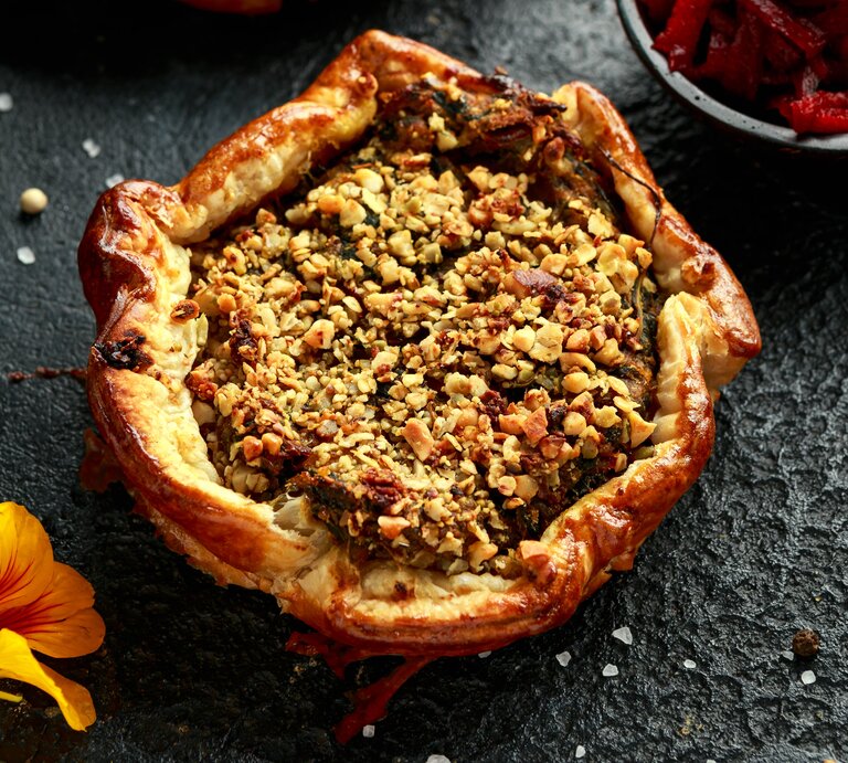 Eine Kürbis-Tarte mit Rote-Bete und Spinat garniert mit Walnüssen, Kürbiskernen und Fetakäse, gold-braun gebacken, neben einer Schüssel Rote-Bete und einer gelben Blume, vor dunklem Hintergrund. | © Getty Images/ DronG