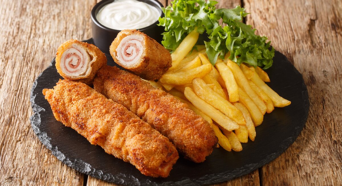 Flamenquín mit Pommes, Salat und Mayonnaise auf Schieferplatte, positioniert auf hölzernem Untergrund. 