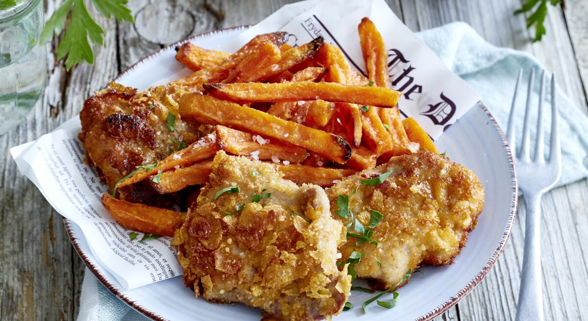 Crisp-Schnitzel mit Süßkartoffelpommes auf einem Teller. Rechts eine Gabel, links davon Petersilie. Draufsicht.
