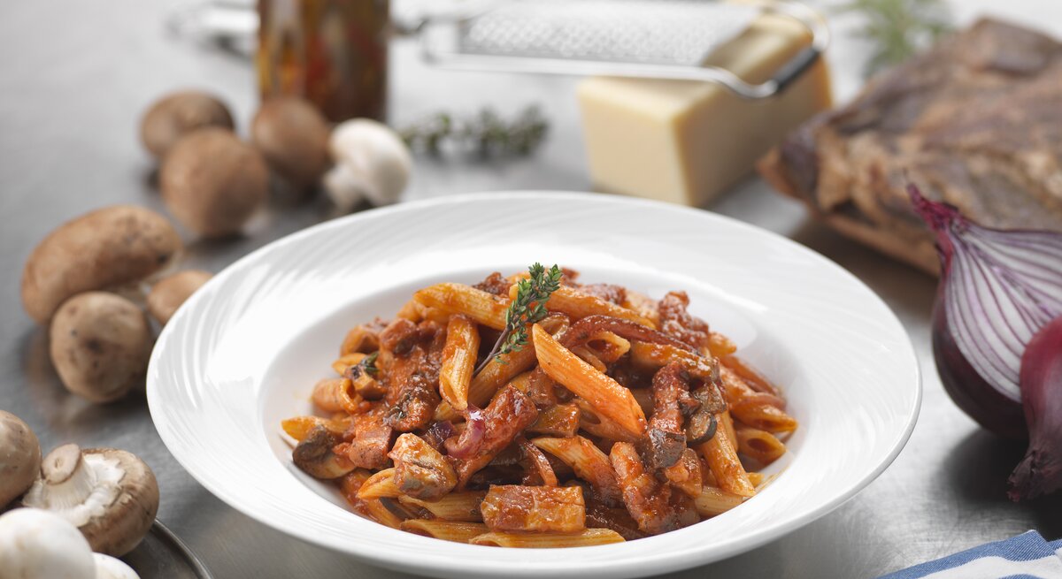 Penne mit Pilzen, Speck und Tomatensoße auf einem weißen Teller.