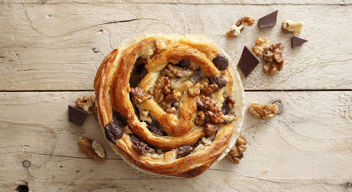 Auf einer Arbeitsplatte aus Holz liegt eine der Blätterteigschnecken mit Schokolade und Walnüssen. Sie ist golbraun gebacken und mit Walnüssen und Schokolade belegt. 