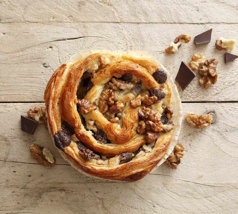 Auf einer Arbeitsplatte aus Holz liegt eine der Blätterteigschnecken mit Schokolade und Walnüssen. Sie ist golbraun gebacken und mit Walnüssen und Schokolade belegt.  | © StockFood / Gross, Petr