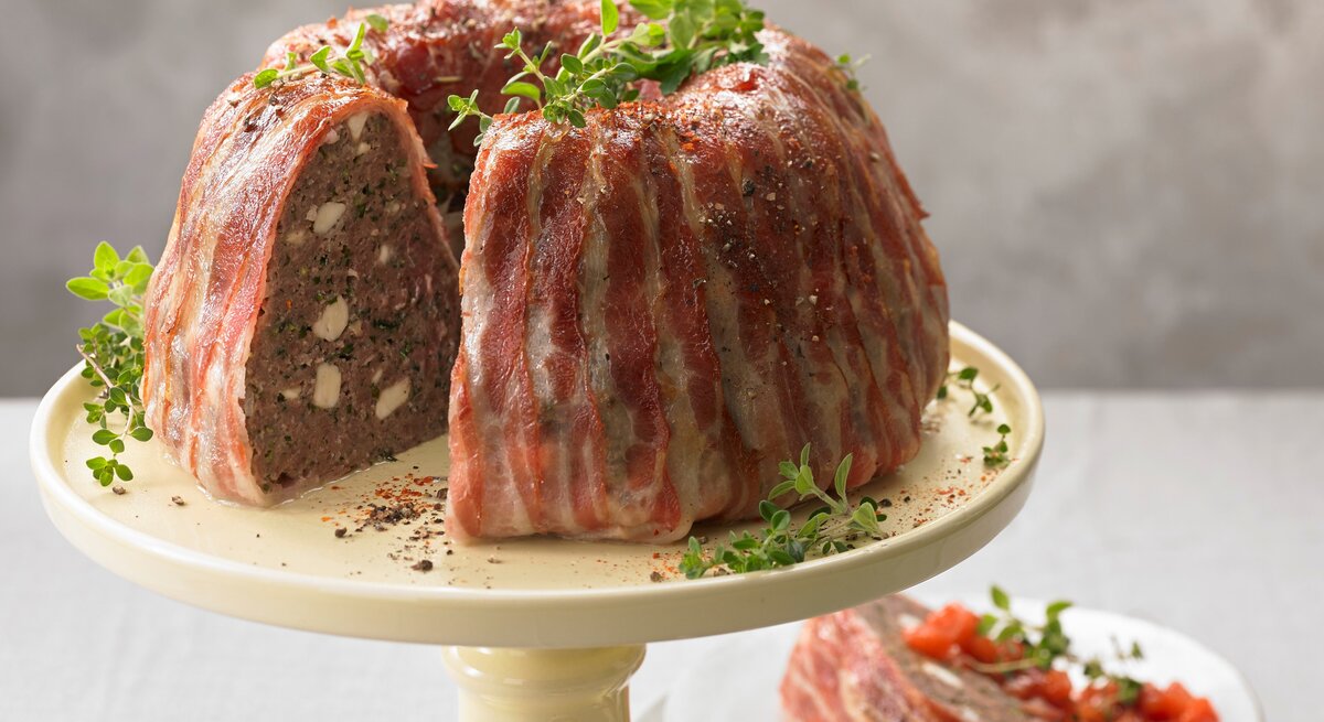Der deftige Gugelhupf mit Rinderhack und Feta im Speckmantel garniert mit Kräutern angerichtet auf einem Kuchenständer vor einem grauen Hintergrund. 