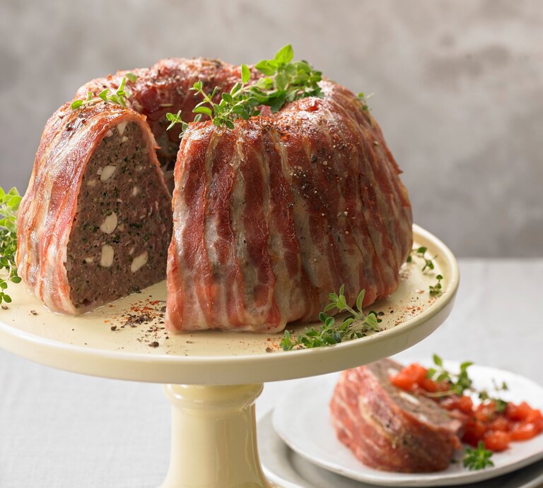 Der deftige Gugelhupf mit Rinderhack und Feta im Speckmantel garniert mit Kräutern angerichtet auf einem Kuchenständer vor einem grauen Hintergrund.  | © StockFood Studios / Westermann, Jan-Peter