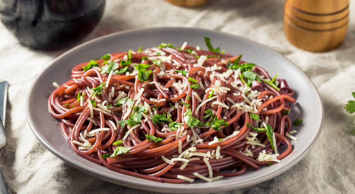 Auf einem Tisch steht ein Teller mit Rotwein-Spaghetti. Daneben sind halb zu sehen: Eine Karaffe mit Rotwein sowie ein Salz- und Pfefferstreuer.