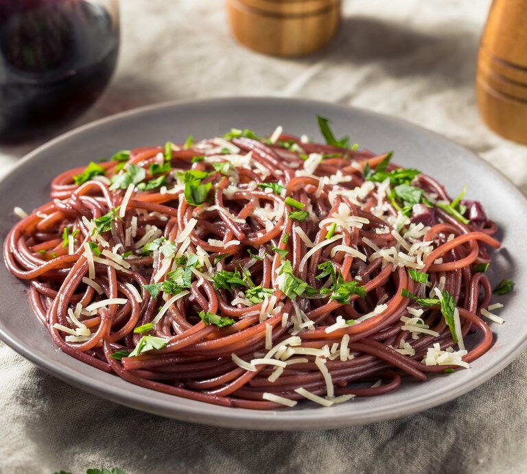 Auf einem Tisch steht ein Teller mit Rotwein-Spaghetti. Daneben sind halb zu sehen: Eine Karaffe mit Rotwein sowie ein Salz- und Pfefferstreuer. | © Getty Images / bhofack2