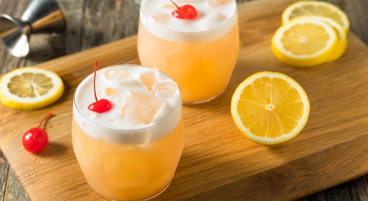 Zwei Raki Sour mit Cocktailkirsche auf Holzbrett auf Holztisch. Zitronenscheiben als Dekoration auf dem Brett verteilt. Liegendes Barmaß am oberen Bildrand. Aufnahme aus der Draufsicht.