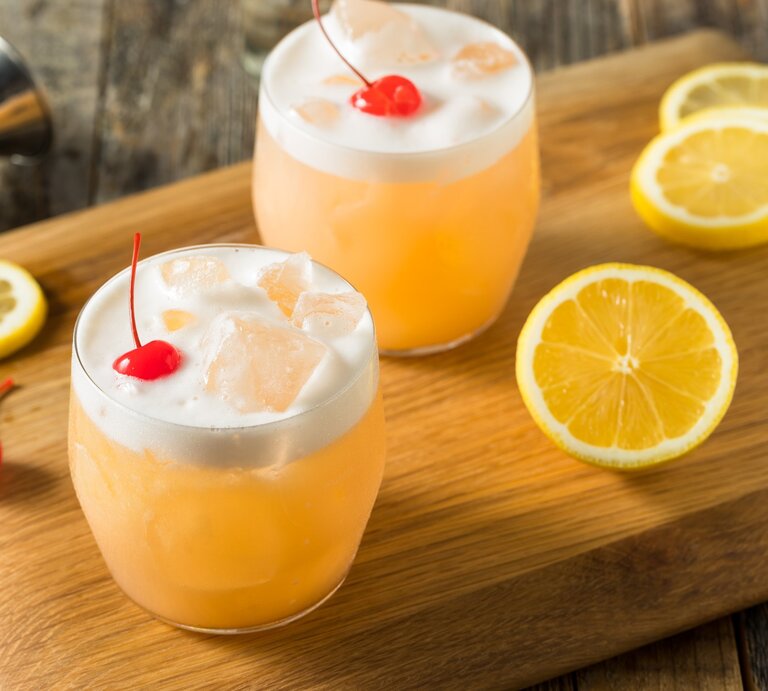 Zwei Raki Sour mit Cocktailkirsche auf Holzbrett auf Holztisch. Zitronenscheiben als Dekoration auf dem Brett verteilt. Liegendes Barmaß am oberen Bildrand. Aufnahme aus der Draufsicht. | © Getty Images/bhofack2