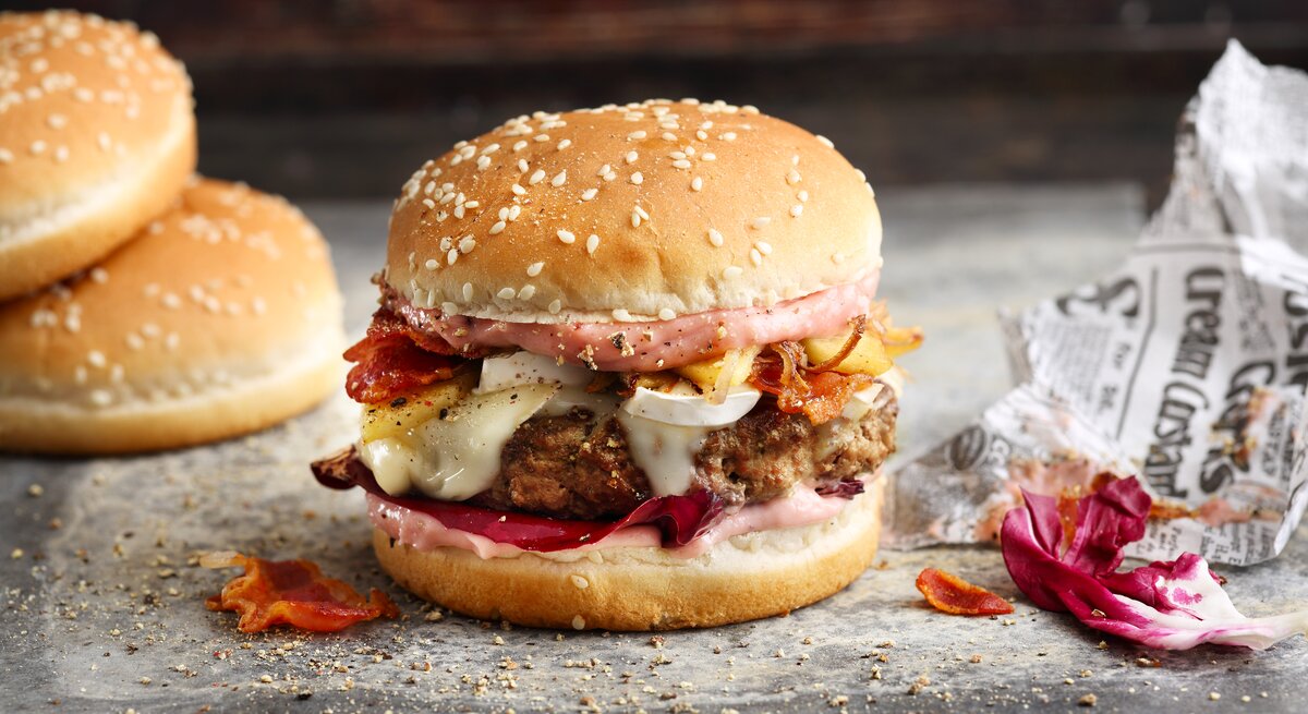 French Burger mit Speck auf einer grauen Steinplatte vor einem dunklen Hintergrund neben Zeitungspapier. 