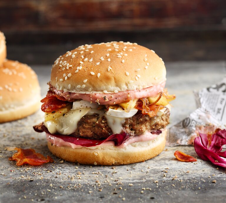 French Burger mit Speck auf einer grauen Steinplatte vor einem dunklen Hintergrund neben Zeitungspapier.  | © Stock Food / Gräfe & Unzer Verlag / Neubauer, Mathias