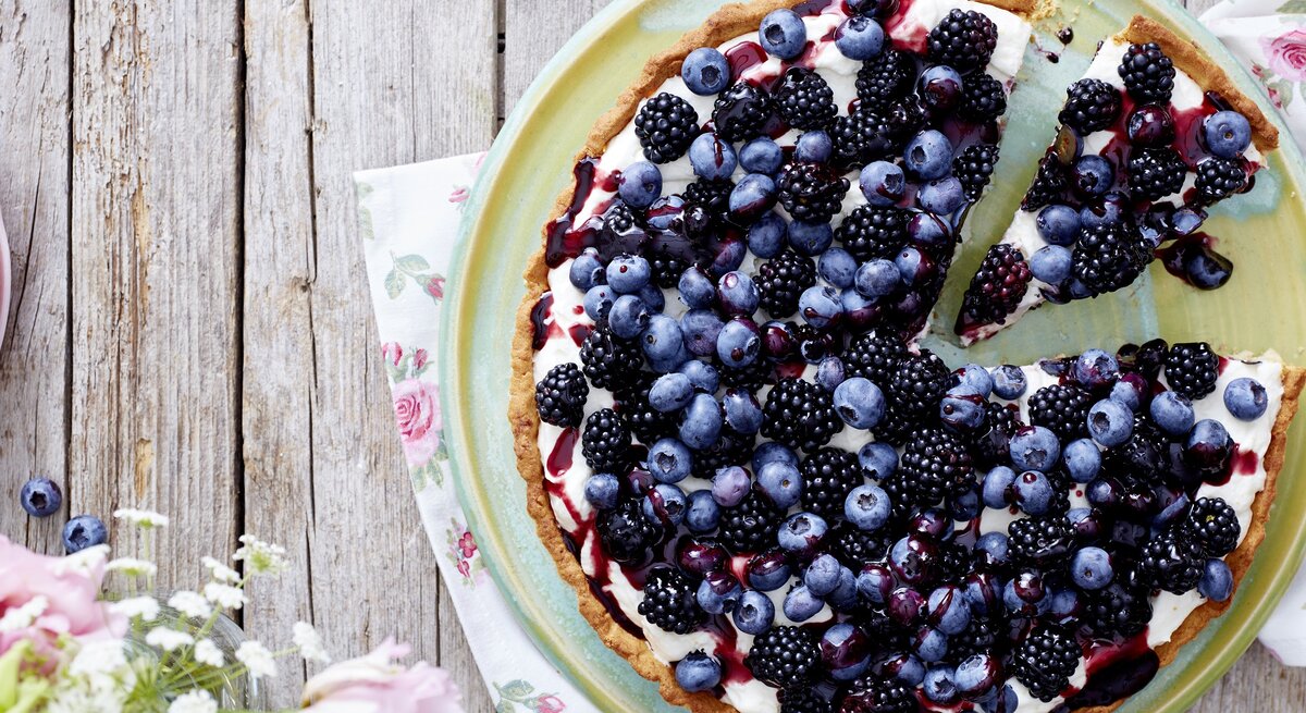 Eine Beeren-Tarte auf einem hellgrünen Teller. Unter dem Teller liegen Servietten mit Blumenmuster. Der Teller steht auf einem Holztisch. Am Bildrand befinden sich Blumen.