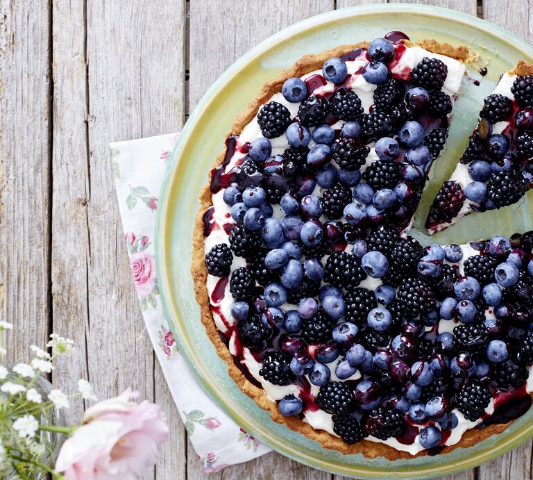 Eine Beeren-Tarte auf einem hellgrünen Teller. Unter dem Teller liegen Servietten mit Blumenmuster. Der Teller steht auf einem Holztisch. Am Bildrand befinden sich Blumen. | © EAT CLUB/Isabell Triemer
