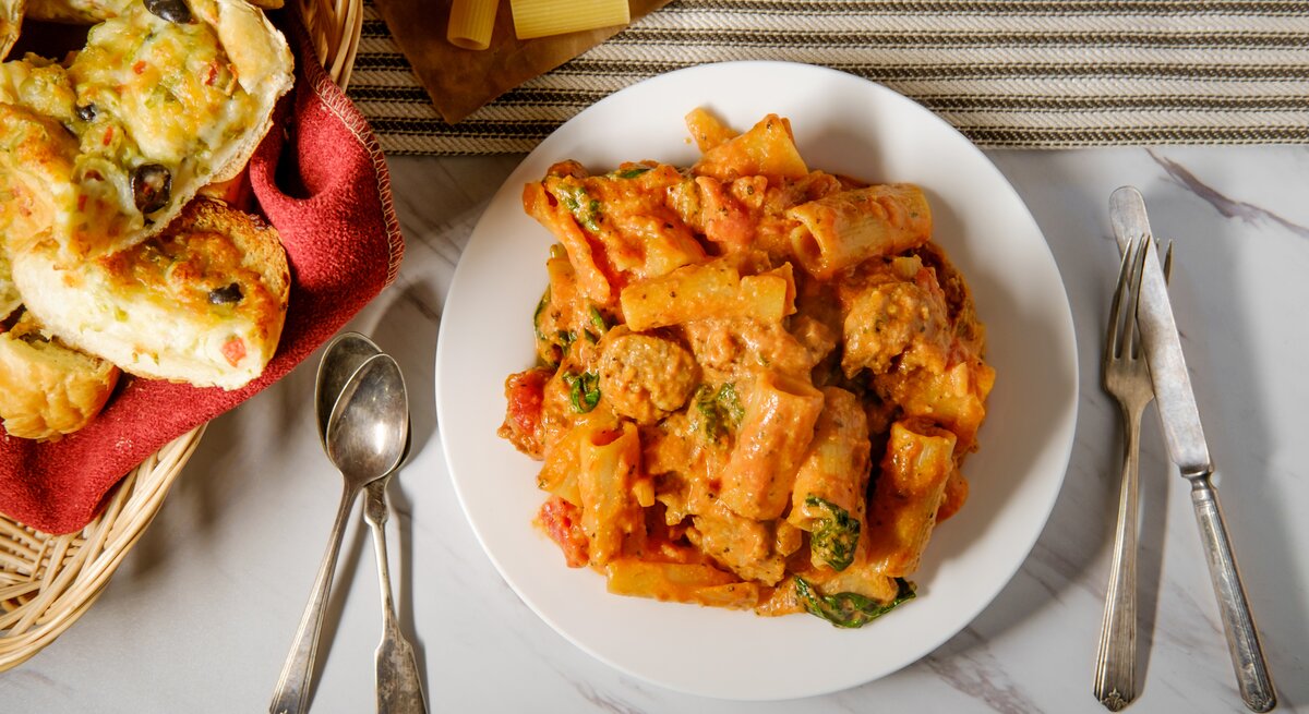 Tortelloni mit Salsiccia in Tomaten-Sahne-Soße auf weißem Teller. Daneben liegen Besteck und selbst gemachtes Knoblauchbrot. 