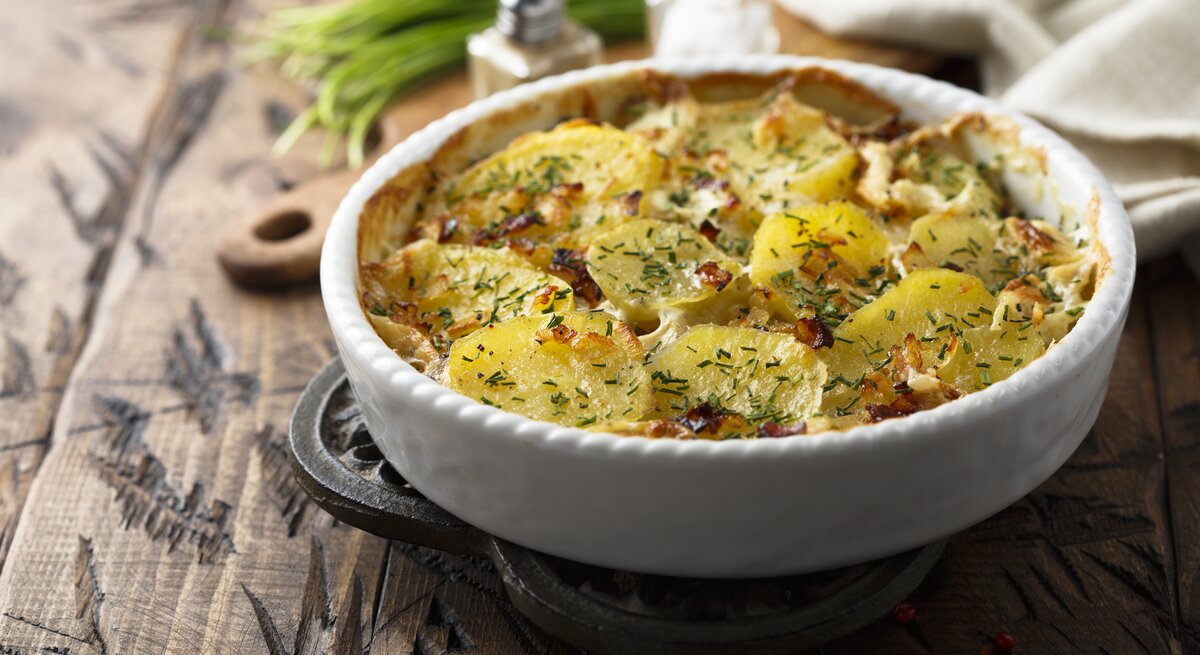 Ein Carbonara-Kartoffelauflauf in einer runden Auflaufform auf einem Holztisch, im Hintergrund unscharf Salz- und Pfefferstreuer, Kräuter, ein Brett und ein Geschirrtuch.