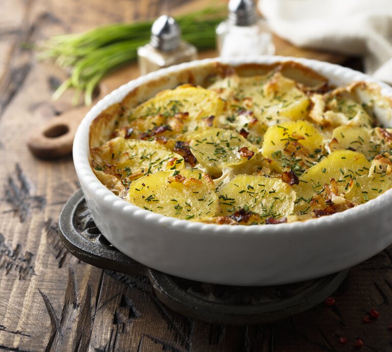 Ein Carbonara-Kartoffelauflauf in einer runden Auflaufform auf einem Holztisch, im Hintergrund unscharf Salz- und Pfefferstreuer, Kräuter, ein Brett und ein Geschirrtuch. | © Getty Images/ Mariha-kitchen