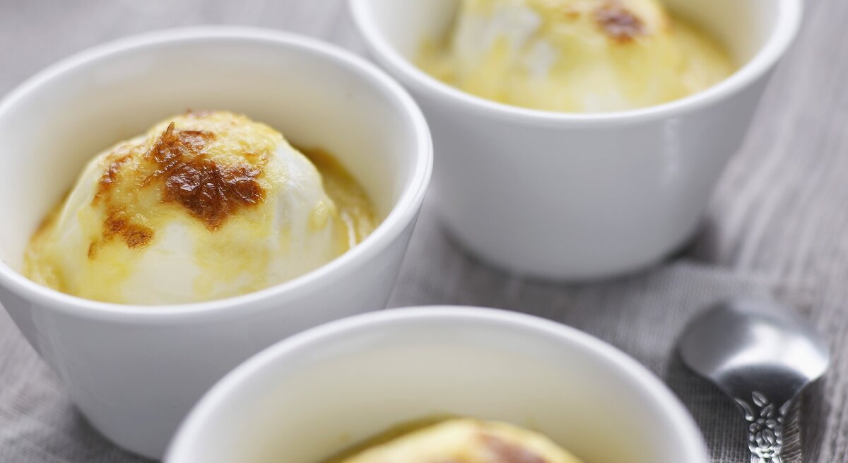 In weißen Förmchen überbackene Eier mit Bechamel, knusprig gratiniert und serviert auf einem grauen Holztisch. 