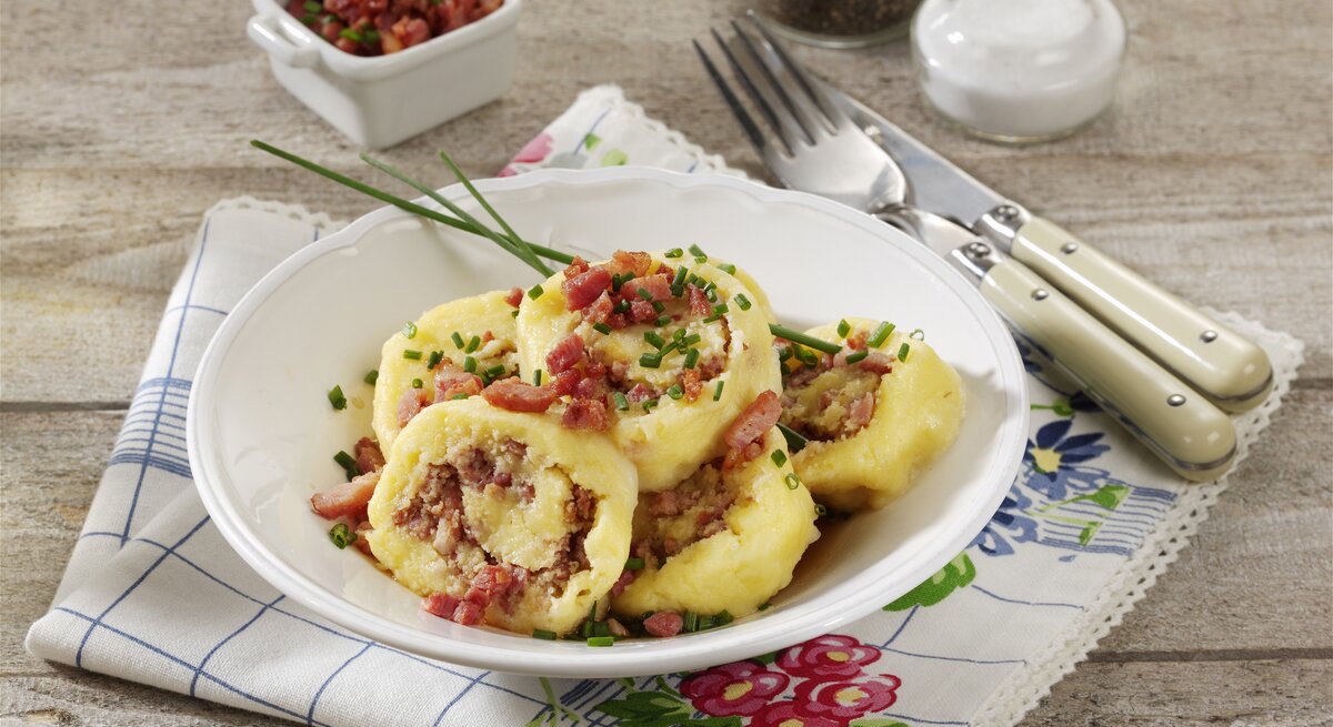 Geschnittene sächsische Wickelknödel auf einem weißen Teller auf einem bedruckten Geschirrhandtuch. Die Wickelknödel sind angerichtet mit Speck und Schnittlauch. Neben dem Teller liegen Messer und Gabel. Im Hintergrund liegt eine Schale mit Speck und zwei Streuer mit Pfeffer und Salz.