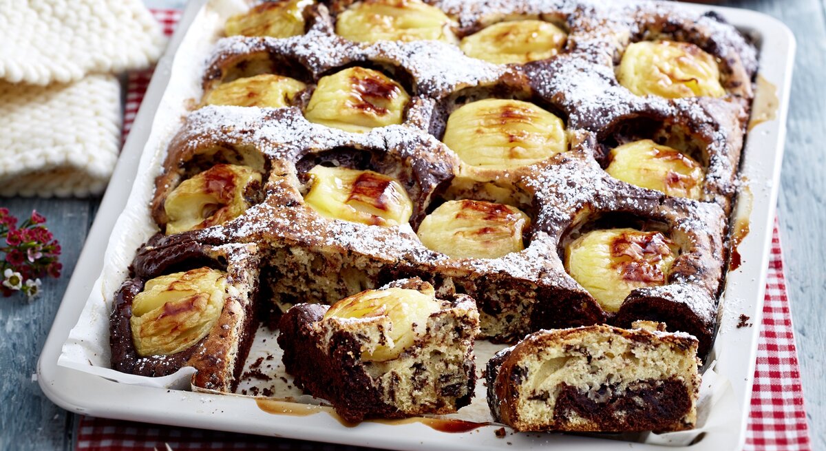 Ein Apfel-Stracciatella-Kuchen mit Gelee und Puderzucker in einem tiefen Blech. Der Kuchen ist angeschnitten und zwei Stücken liegen am Rand der Form.