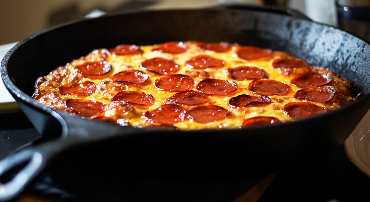 Pizza mit Salami in einer gusseisernen Pfanne. Dahinter verschiedene Behälter. 
