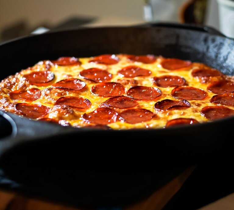 Pizza mit Salami in einer gusseisernen Pfanne. Dahinter verschiedene Behälter.  | © GettyImages/DianeBentleyRaymond