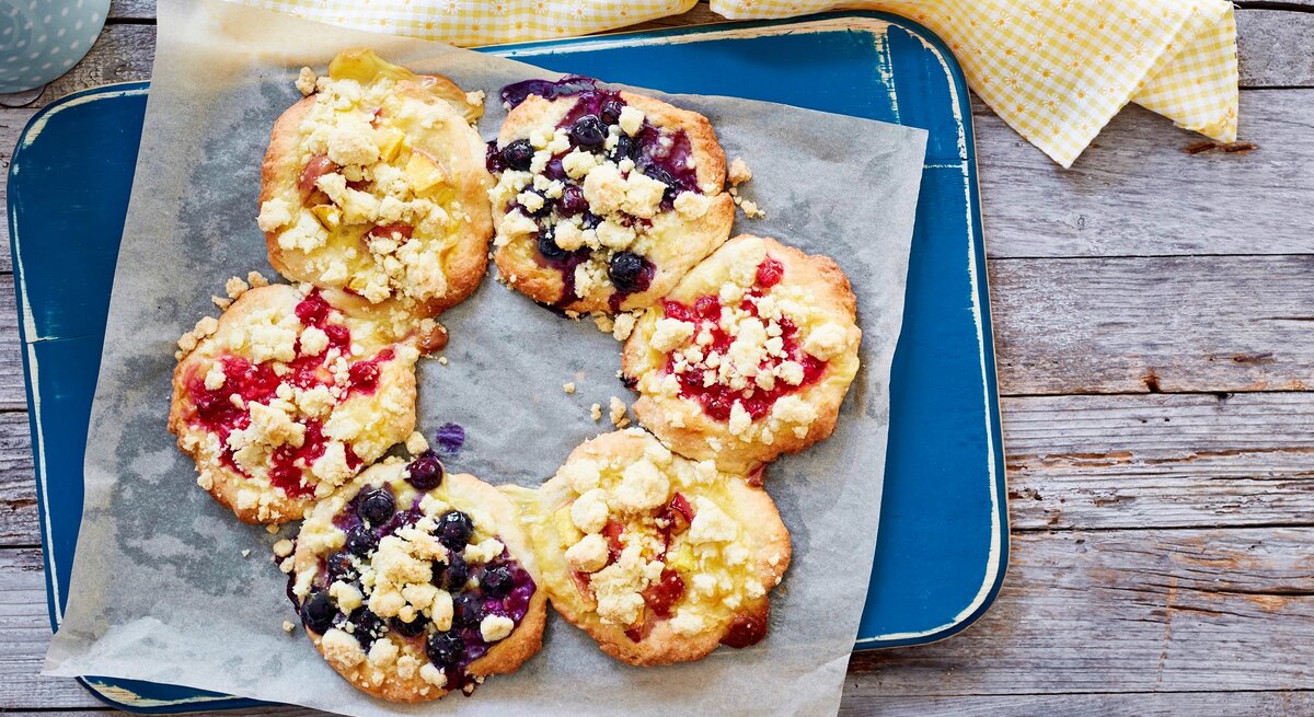 Streuseltaler-Kranz mit fruchtigen Beeren und Vanillepudding, serviert auf einem hellblauen Tablett auf einem groben Holztisch.