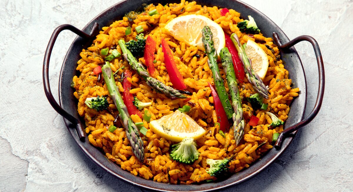 Eine vegane Paella mit grünem Spargel, Zitronenscheiben, Paprika und Brokkoli in einer typischen Paella-Pfanne.