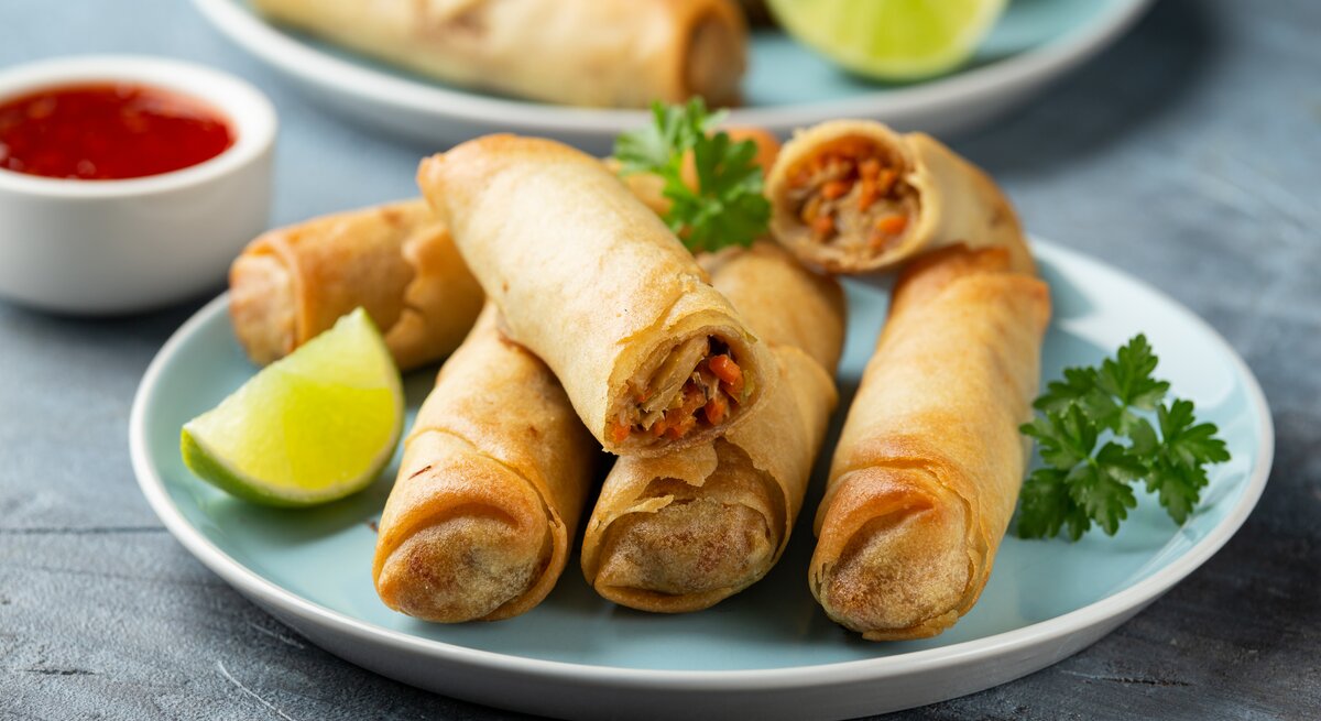 Auf zwei blauen Tellern liegen vegane Frühlingsrollen, dazwischen steht eine weiße Schale mit roter Soße zum Dippen.