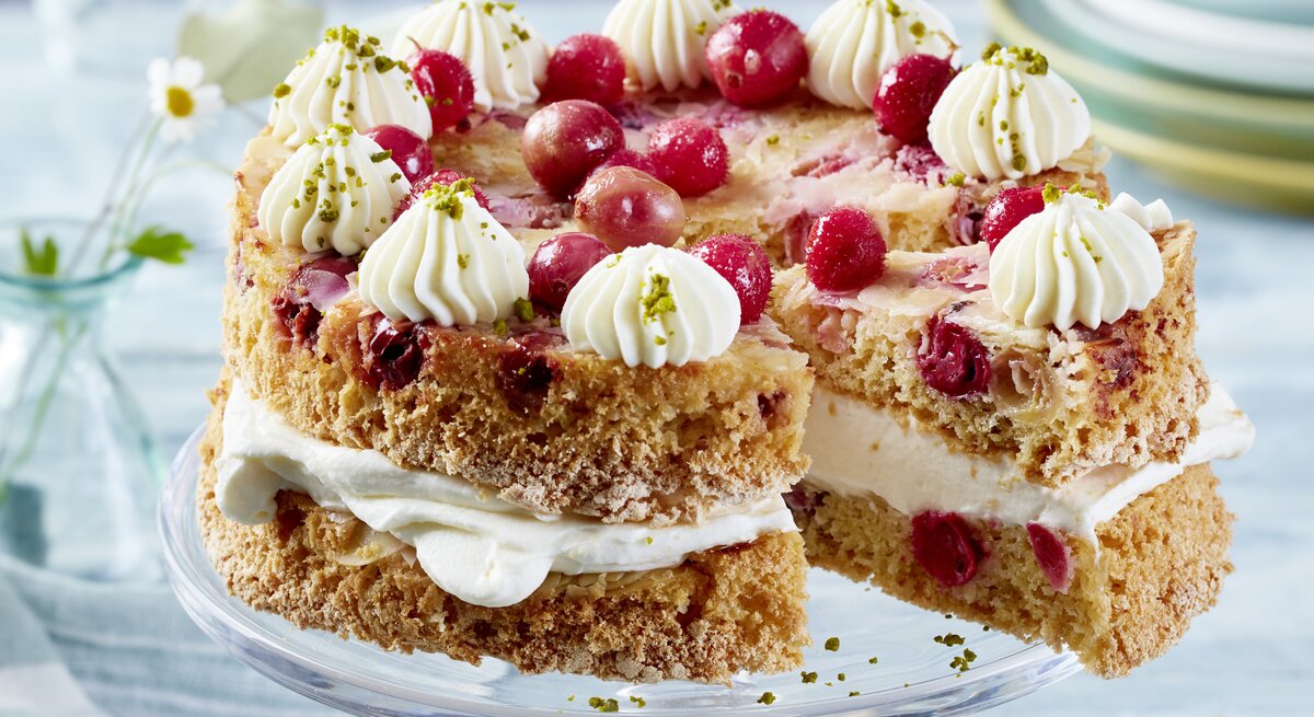 Eine zweilagige Torte mit hellem Teig und roten Johannisbeeren, gefüllt und dekoriert mit weißer Sahnecreme. Die Torte ist auf einem Glas-Tortenständer angerichtet und mit weiteren Johannisbeeren und gehackten Pistazien garniert. Im Hintergrund stehen gestapelte Teller und Glasvasen mit kleinen Blumen.