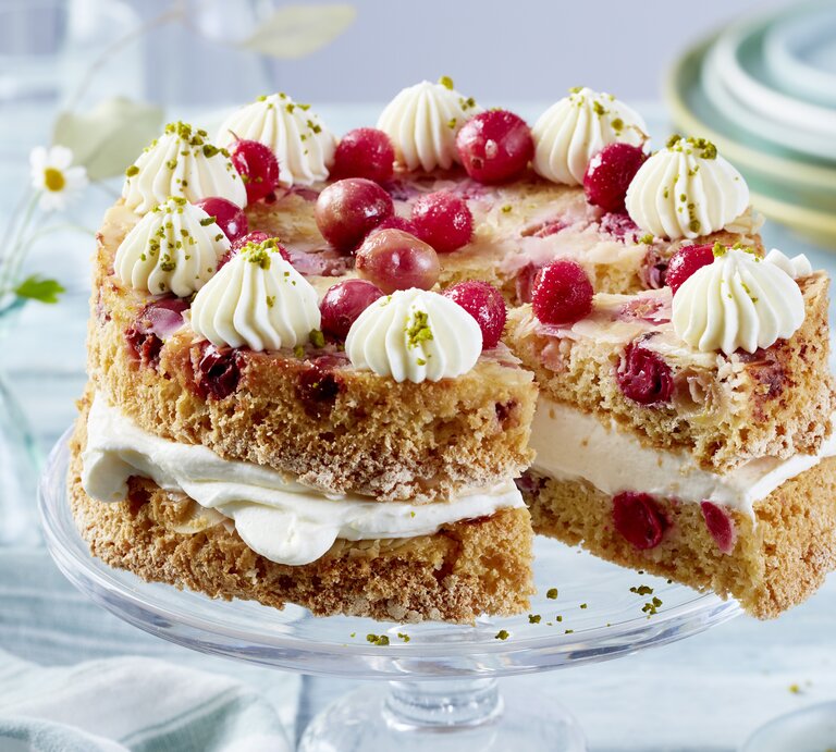 Eine zweilagige Torte mit hellem Teig und roten Johannisbeeren, gefüllt und dekoriert mit weißer Sahnecreme. Die Torte ist auf einem Glas-Tortenständer angerichtet und mit weiteren Johannisbeeren und gehackten Pistazien garniert. Im Hintergrund stehen gestapelte Teller und Glasvasen mit kleinen Blumen. | © EAT CLUB/Isabell Triemer