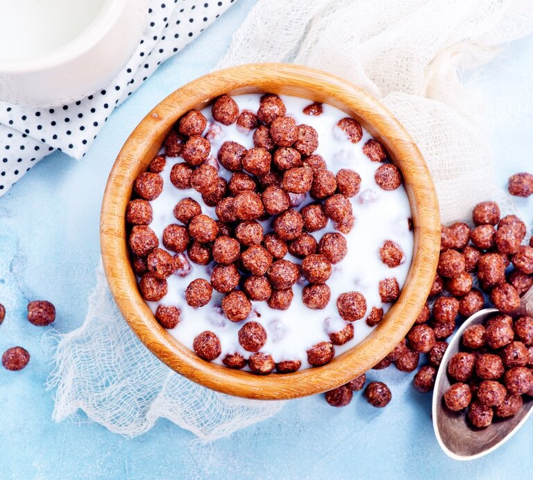 Eine braune Schale mit den knusprigen Schoko-Kugeln und Milch auf hellblauem Untergrund. Daneben ein silberner Löffel und ein weißes Milchkännchen. | © Shutterstock/ Gayvoronskaya_Yana